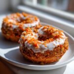 Two glazed Vegan Carrot Cake Olive Oil Donuts topped with shredded carrots on a white plate.