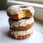 A stack of three Vegan Lemon Poppy Seed Donuts, topped with a thick, speckled lemon glaze. The top donut has a bite taken out.
