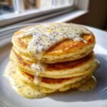 Close-up of a stack of fluffy Vegan Lemon Poppyseed Pancakes topped with a thick, creamy lemon glaze dotted with poppy seeds.
