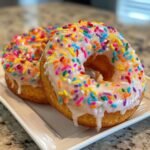 Two fluffy Vegan Vanilla Donuts With Sprinkles, covered in white glaze and colorful jimmies, resting on a white plate.