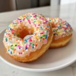 Two fluffy Vegan Vanilla Donuts With Sprinkles, featuring a glossy vanilla glaze and colorful rainbow sprinkles.