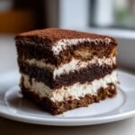 A perfect slice of Delicious Coffee Tiramisu Cake Recipe with layers of dark cake, cream, and cocoa dusting.