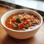 A steaming white bowl filled with Flavorful Stuffed Pepper Soup, featuring ground meat, rice, and chunks of red and green bell peppers.