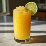 A tall glass filled with a bright yellow, icy Frozen Pineapple Margarita Mocktail, garnished with a fresh lime wheel.