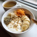 A white bowl filled with yogurt, topped with banana slices, seeds, sprouts, grated carrots, and grains, representing foods to improve gut health.