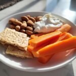 A plate showing components for healthy snacking tips: carrot sticks, whole almonds, crackers, and a dollop of yogurt dip.