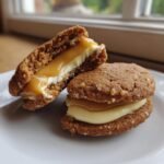 Two Irresistible Apple Cider Whoopie Pie Cookies filled with cream and gooey caramel, one cut in half.