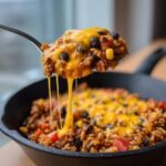 A spoonful of Irresistible Cheesy Taco Skillet With Rice Recipe showing a dramatic cheese pull over the skillet.
