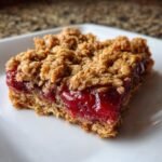 A single, thick square of Irresistible Cherry Crumble Bars showing a bright red cherry filling between an oat base and crumble topping.