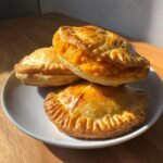 Three golden-brown Halloween Hand Pies Bites stacked on a white plate, showing a bright orange filling spilling out.