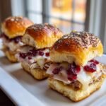 Three Irresistible Havarti Cranberry Turkey Sliders lined up on a white platter, featuring melted cheese and cranberry sauce.