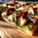Close-up of a slice of Irresistible Pesto Chicken Flatbread topped with melted cheese, pesto, and sun-dried tomatoes.