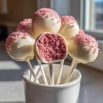 Close-up of Irresistible Strawberry Cake Pops dipped in white chocolate and pink sprinkles, one is bitten to show the pink cake center.