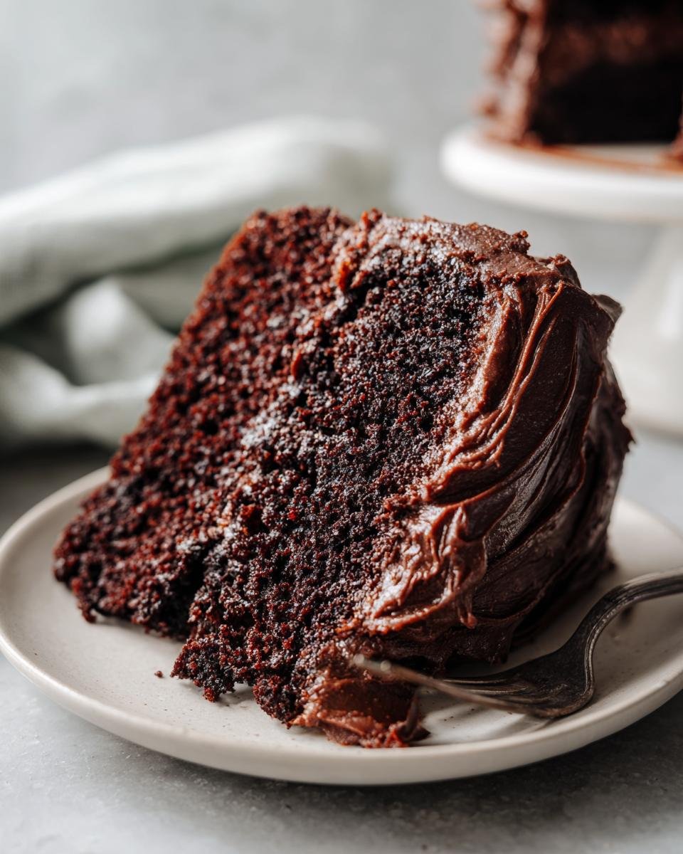 Close-up of a moist slice of dark cake, likely Irresistible Gingerbread Cake, covered in rich chocolate frosting with a fork taking a bite.