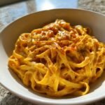 Close-up of creamy fettuccine coated in bright orange sauce, seasoned with black pepper and red pepper flakes, representing One Pot Cajun Pumpkin Alfredo Pasta.