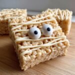 A close-up of a Spooktacular Halloween Mummy Rice Krispie Treat decorated with white icing bandages and candy eyes.