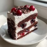 A perfect slice of Black Forest Cake showing layers of dark chocolate sponge, white cream, cherries, and chocolate shavings.
