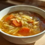 Close-up of a steaming white bowl filled with Cozy Cabbage Soup Recipe, featuring shredded cabbage and sliced carrots in a rich broth.