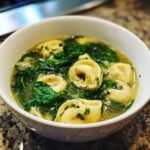 A close-up bowl of Cozy Spinach And Tortellini Soup, featuring pasta and wilted greens in broth.
