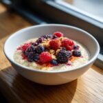 A white bowl filled with creamy quinoa porridge topped generously with fresh raspberries, blueberries, blackberries, and granola.