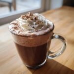 A close-up of Decadent Crockpot Hot Chocolate topped with whipped cream and cocoa powder in a clear mug.