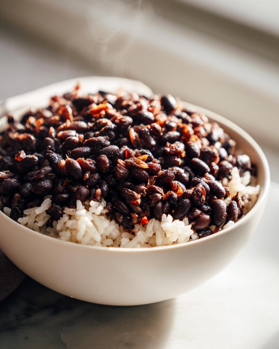 Close-up of a white bowl filled with white rice topped generously with steaming black beans for a Delicious Rice And Beans recipe.