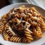 Close-up of rotini pasta tossed in rich caramelized onions, topped with melted white cheese for a delicious French Onion Pasta Recipe.