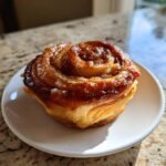 A single, golden-brown, glazed Peach Pie Crescent Cruffin sitting on a small white plate.