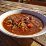 A close-up of a rich, dark red Hearty Hobo Stew filled with ground meat, carrots, and beans, served in a white bowl outdoors.