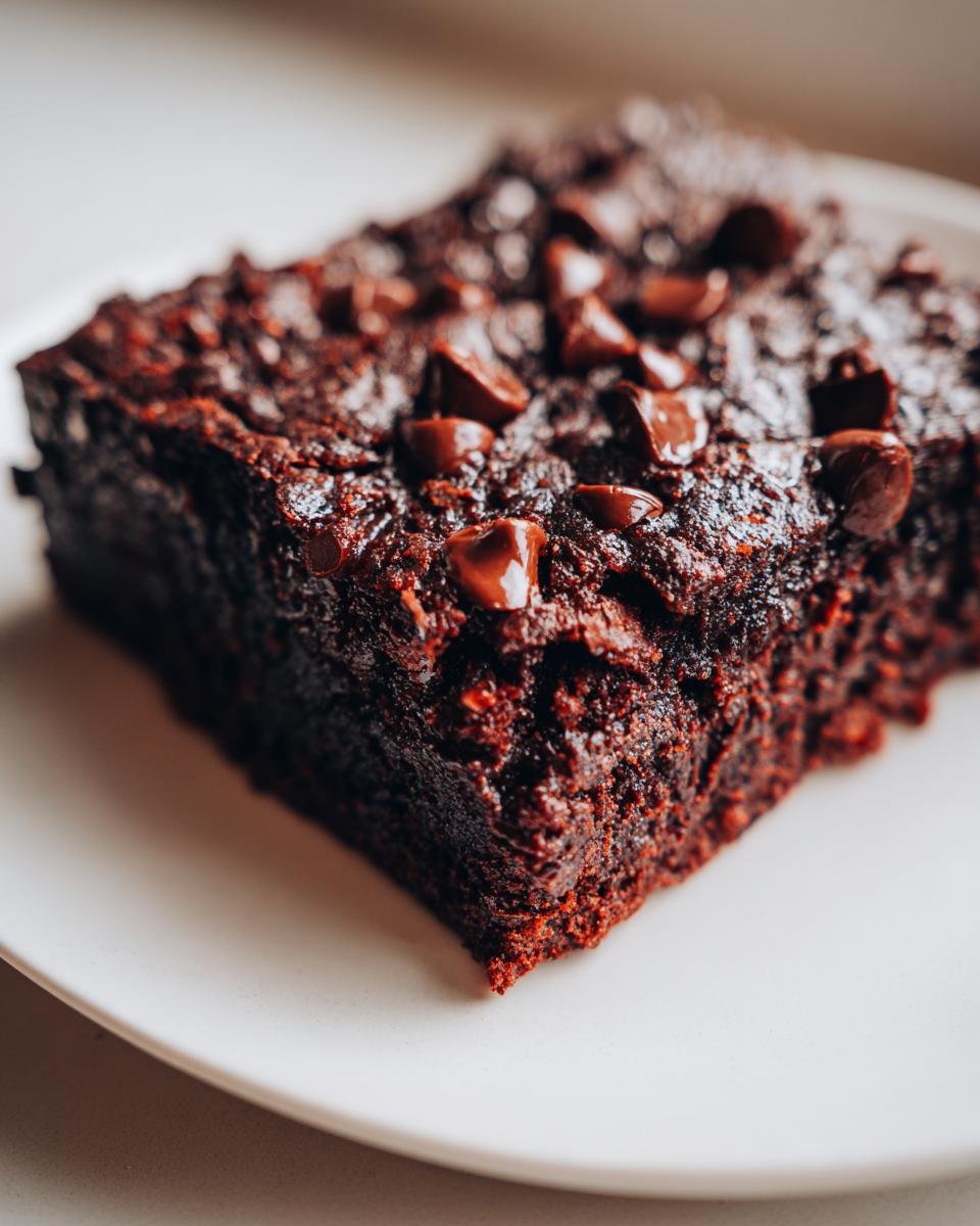 A rich, dark square of Indulgent Sweet Potato Brownies topped with melted chocolate chips on a white plate.