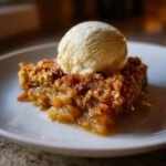 A warm slice of Irresistible Apple Crisp Recipe topped with a scoop of melting vanilla ice cream on a white plate.