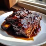 Close-up of tender, glazed Irresistible Balsamic Bourbon Short Ribs resting in their rich sauce on a white plate.