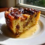A moist slice of Irresistible Banana Bread Pudding, studded with raisins and topped with a caramelized crust, sitting in a pool of sauce.