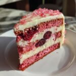 A cross-section slice of Irresistible Cherry Layer Cake With Cream Cheese Frosting showing pink layers, white frosting, and whole cherries.