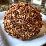 A close-up of an Irresistible Chipped Beef Cheeseball completely coated in chopped pecans, served on a white square plate.