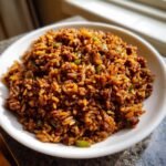 A close-up of a white bowl filled with rich, dark brown Irresistible Dirty Rice Recipe mixed with ground meat and green peppers.
