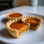 Three Irresistible Mini Pumpkin Pies on a white plate, one pie is cut in half showing the rich orange filling.