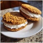 Two golden brown Irresistible Oatmeal Cream Pies filled with thick white cream filling, resting on a white plate.