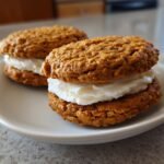 Two homemade Irresistible Oatmeal Cream Pies stacked with thick white frosting, served on a white plate.