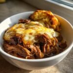 A bowl of shredded beef topped with melted cheese and onions, served with a biscuit, showcasing the Irresistible Savory French Onion Pot Roast.