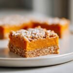 A close-up of one square of Irresistible Spiced Pumpkin Pretzel Bars, showing the thick orange pumpkin layer and oat crust, dusted with powdered sugar.