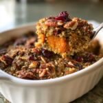 A spoonful of Irresistible Sweet Potato Quinoa Casserole topped with pecans and cranberries being lifted from a white baking dish.