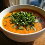 A close-up of a white bowl filled with vibrant orange Irresistible Thai Coconut Red Lentil Soup, topped generously with fresh chopped cilantro.