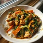 A close-up of Irresistible Thai Peanut Chicken Stir Fry with chicken, carrots, and greens served over white rice in a white bowl.