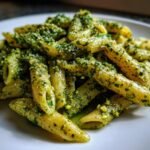 Close-up of penne pasta coated in vibrant green pistachio pesto, served on a white plate.