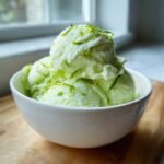 Three scoops of light green, icy Refreshing Pickle Margarita Sorbet topped with bright green lime zest in a white bowl.