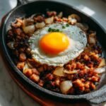 A close-up of Sizzling Sisig Matua served on a hot cast iron plate, topped with a perfectly cooked sunny-side-up egg and onions.
