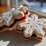 Two Irresistible Snowflake Linzer Cookies filled with bright red raspberry jam and dusted heavily with powdered sugar.