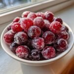 Close-up of bright red Sparkling Sugared Cranberries piled high in a small white bowl near a window.
